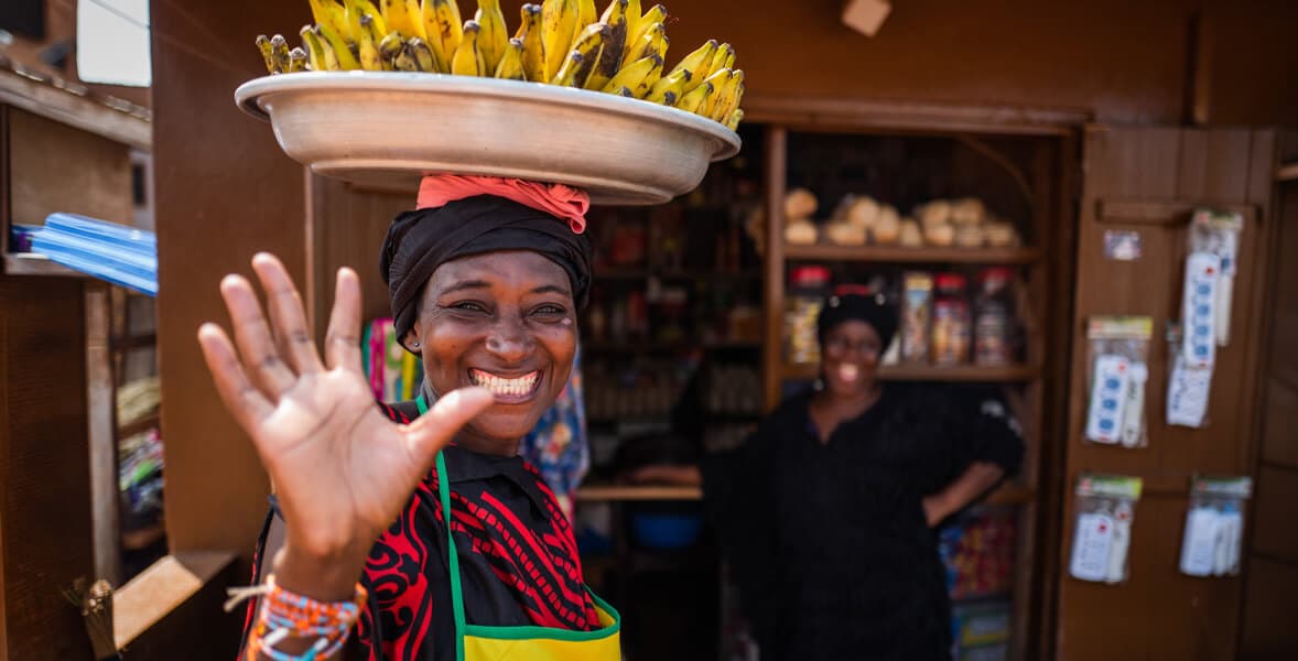 Vendor waving at a local market with a tray of bananas balanced on her head.